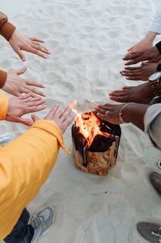 A diverse group warming their hands around a beach bonfire on sandy ground.