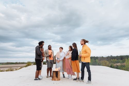 Free A diverse group of friends gathering around a bonfire on a sandy beach under cloudy skies. Stock Photo