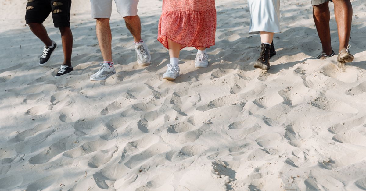 A diverse group of people walking on a sandy beach, showcasing summer fashion and casual footwear.
