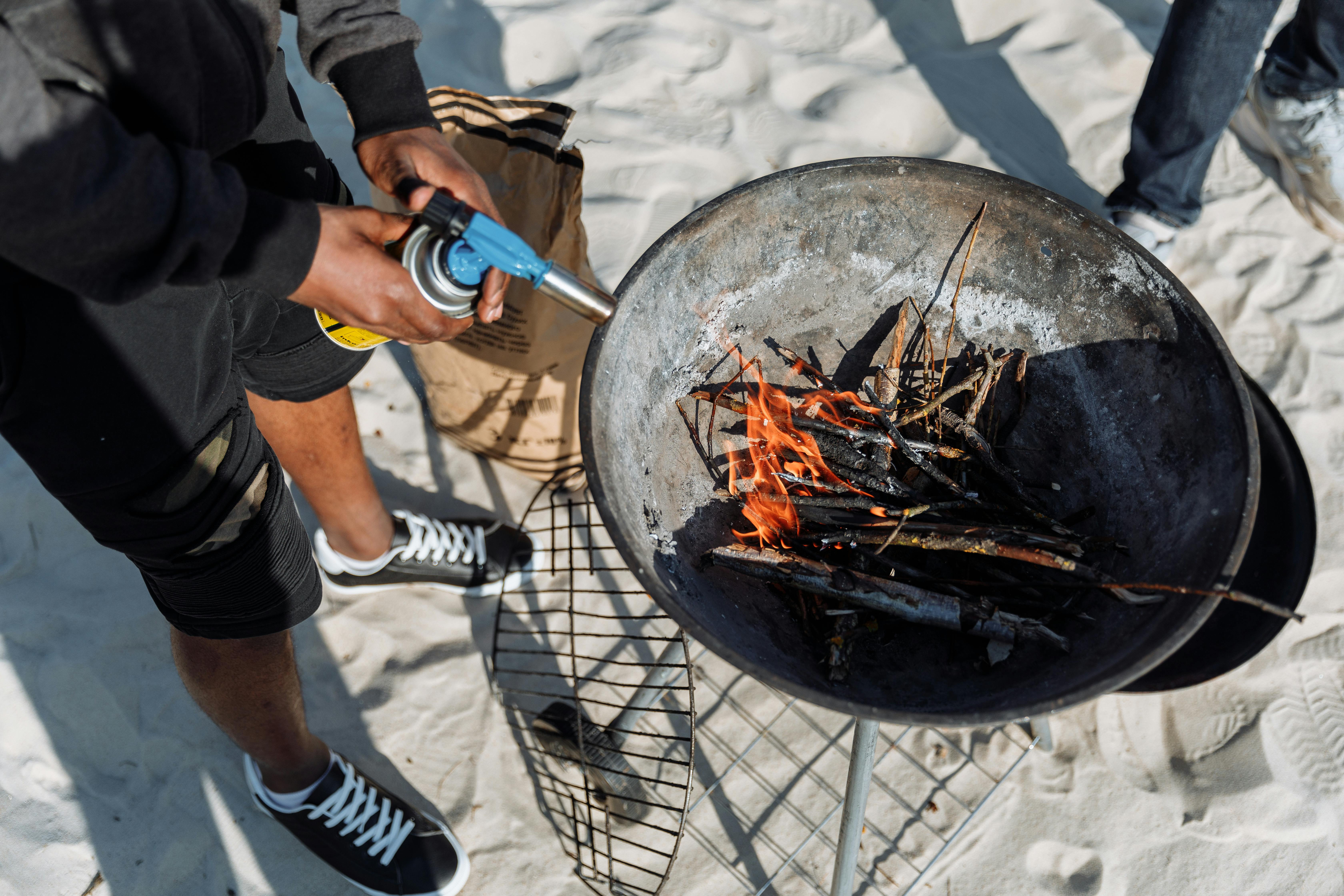 Close-up of person using a grill torch to light fire for outdoor cooking on the beach.