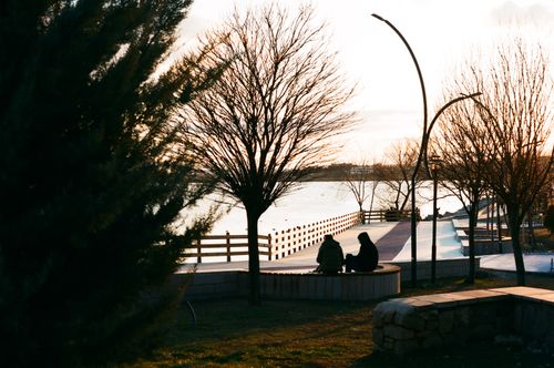 Free Two individuals sit together in a park, silhouetted against a serene lakeside sunset. Stock Photo