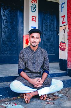 Confident young man in traditional outfit sitting outdoors with colorful backdrop.