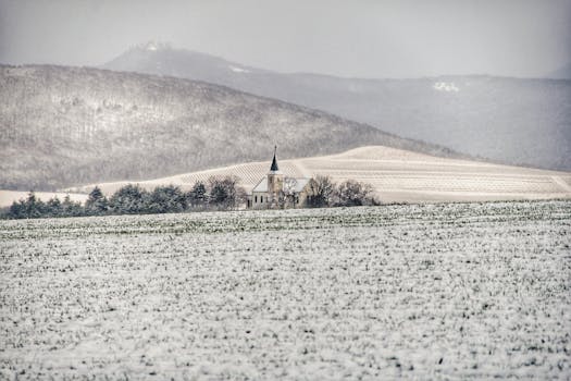 冬日宁静的景色中，教堂与白雪覆盖的田野相映成趣，背景是雾气缭绕的山脉