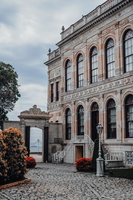A detailed view of a historic palace facade in İstanbul, showcasing ornate architecture with stairs, windows, and lamp posts.