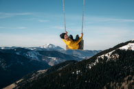 Back view of young male traveler swinging on wooden swing against picturesque landscape of snowy mountains cover with trees and looking at camera over shoulder under blue sky