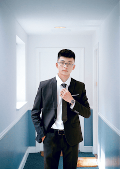 Asian man in suit with eyeglasses in a bright hallway, looking confident.