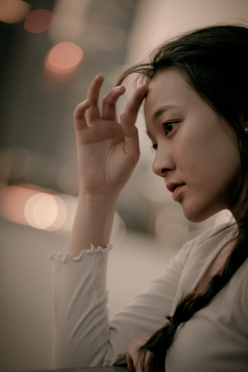 Free A young East Asian woman in thoughtful pose, evening city lights in the background. Stock Photo