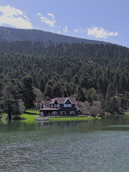 A picturesque house beside a tranquil lake with lush forests and towering mountains in the background.