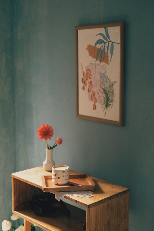 Wooden cabinet with mug on tray near vase with flowers near painting on wall in light apartment