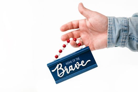 Close-up of a hand holding a "Home of the Brave" decorative sign against a white background.