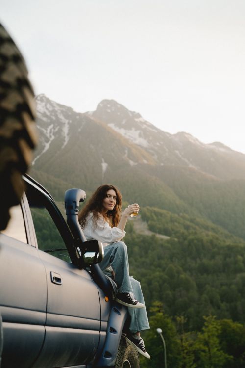 Free Woman sitting on SUV enjoying a scenic mountain view during the day, representing travel and adventure. Stock Photo