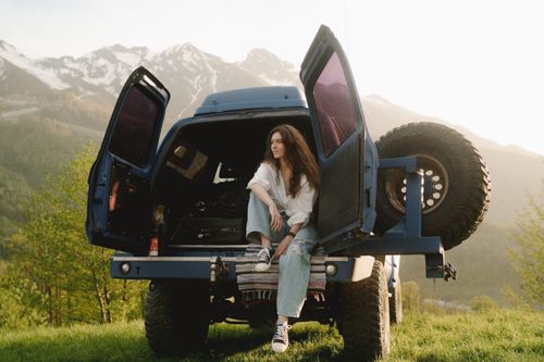Free Young woman enjoys a scenic mountain view sitting in a 4x4 vehicle at sunset. Stock Photo
