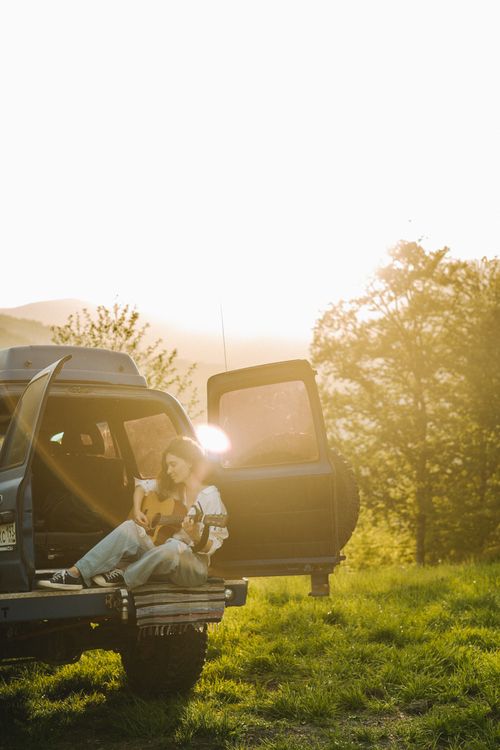 Free A woman sits on an SUV with a guitar in a sunlit countryside setting, embodying freedom and music. Stock Photo