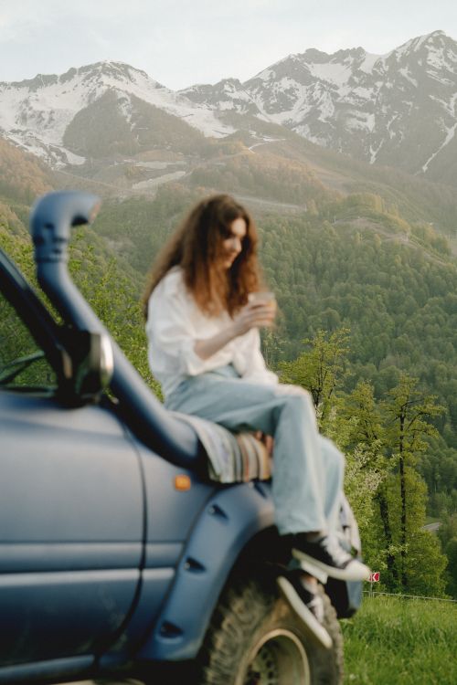 Free Woman enjoying a peaceful moment sitting on a vehicle against a scenic mountain backdrop. Stock Photo