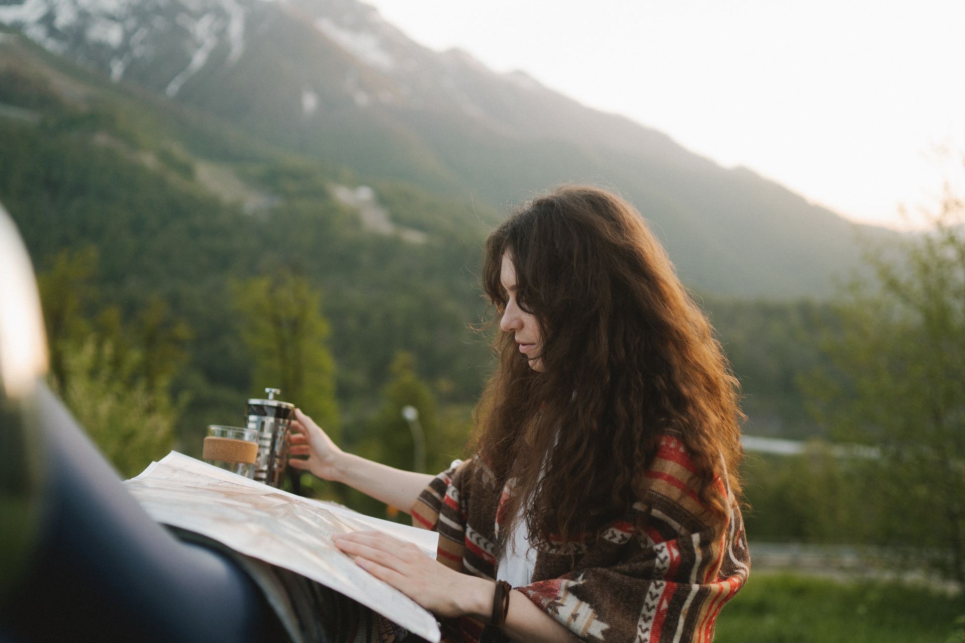 Brunette woman with map relaxing outdoors with coffee in a mountainous setting during sunset.