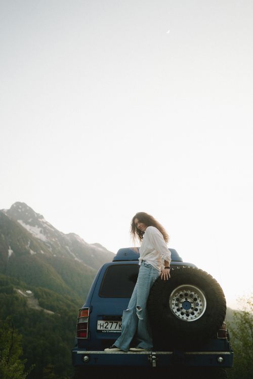 Free A woman stands on a car in a scenic mountain setting during sunset, embracing nature. Stock Photo