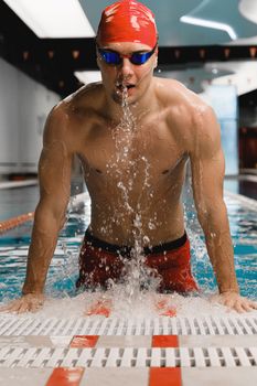 Athletic man in goggles and swim cap emerging from an indoor pool with water splashes.