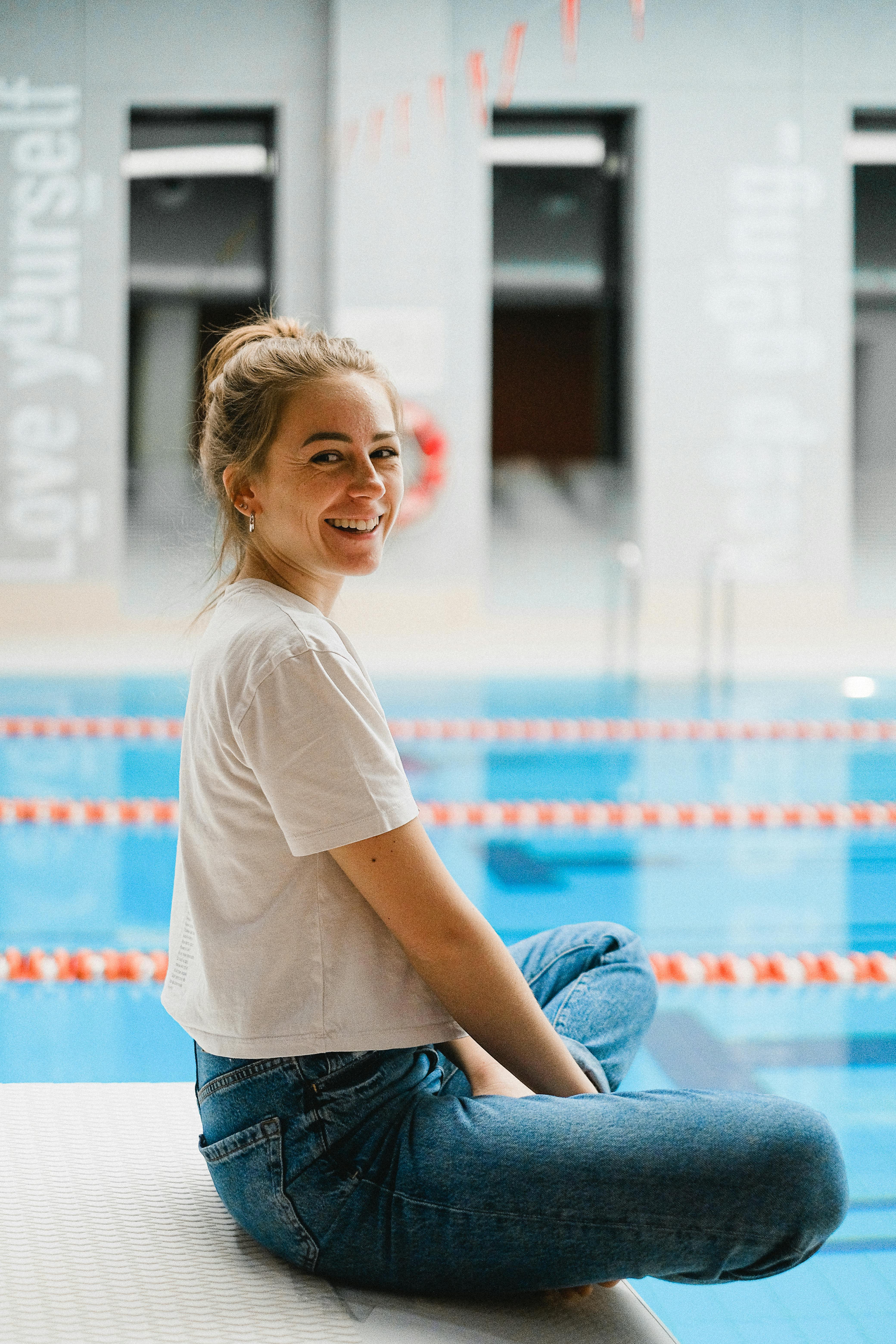 Casual woman in denim sitting by an indoor pool, smiling softly with a relaxed demeanor.
