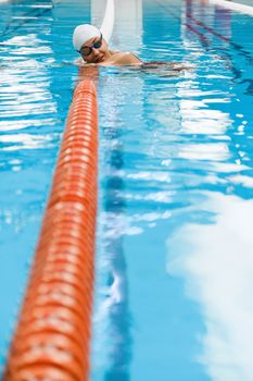 Female swimmer in white cap and goggles relaxing by lane rope in an indoor pool.