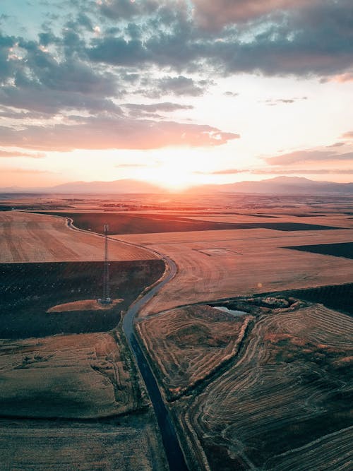 Drone view of endless field and narrow curvy road under cloudy sky in sunset