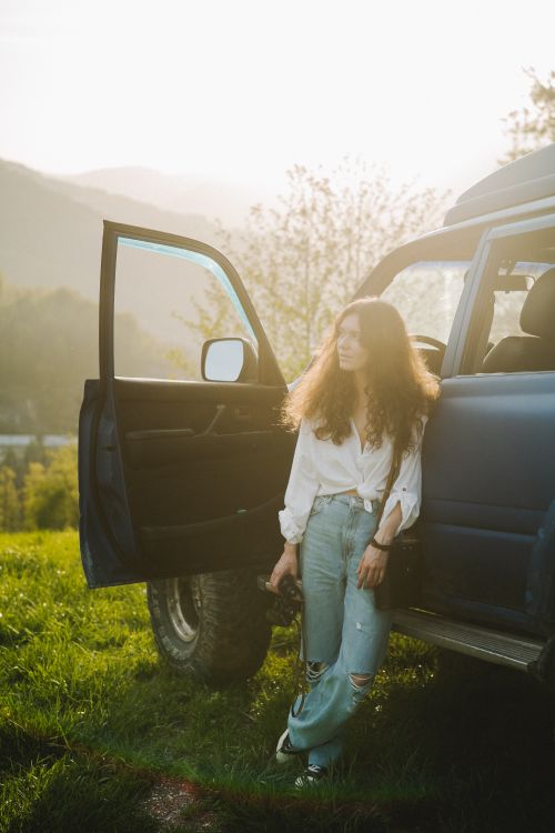 Free Woman with curly hair stands by a car in the countryside at sunset. Stock Photo