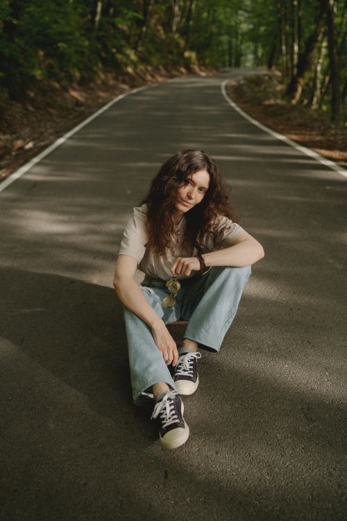 Free Casual young woman sitting on an empty rural road surrounded by greenery in the summer. Stock Photo
