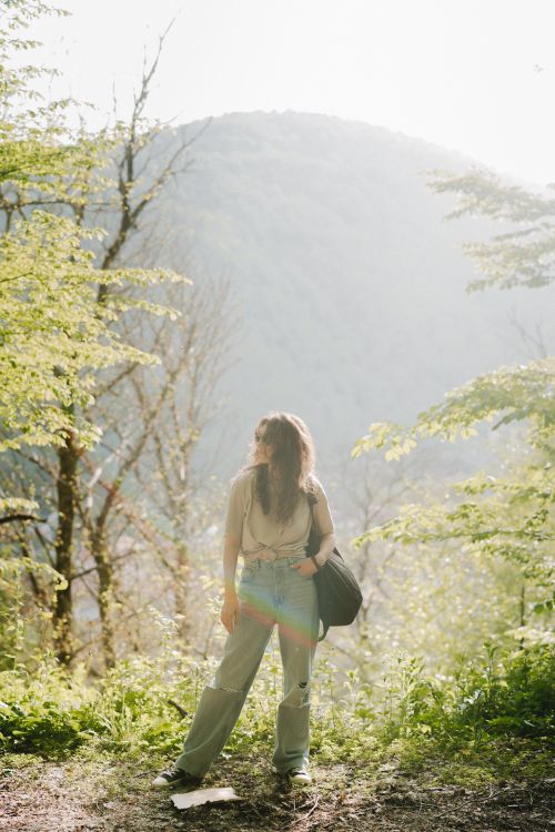 Free Woman standing in vibrant forest, basking in sunlight with scenic mountain in the background. Stock Photo