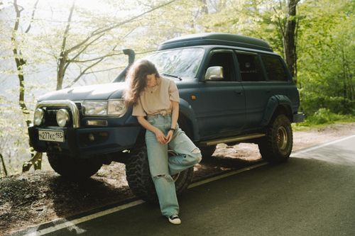 Free A young woman leans on an SUV, enjoying the serene mountain road setting in ripped jeans and a casual t-shirt. Stock Photo