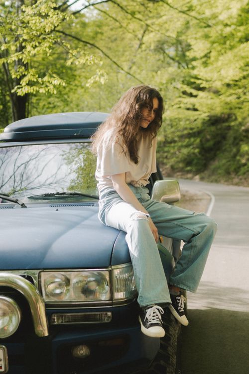 Free Fashionable woman sitting on a car hood, blending urban style with nature's tranquility. Stock Photo