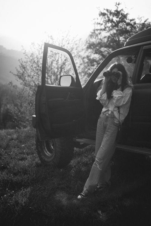 Free Black and white photo of a woman with binoculars by an off-road vehicle, capturing exploration. Stock Photo