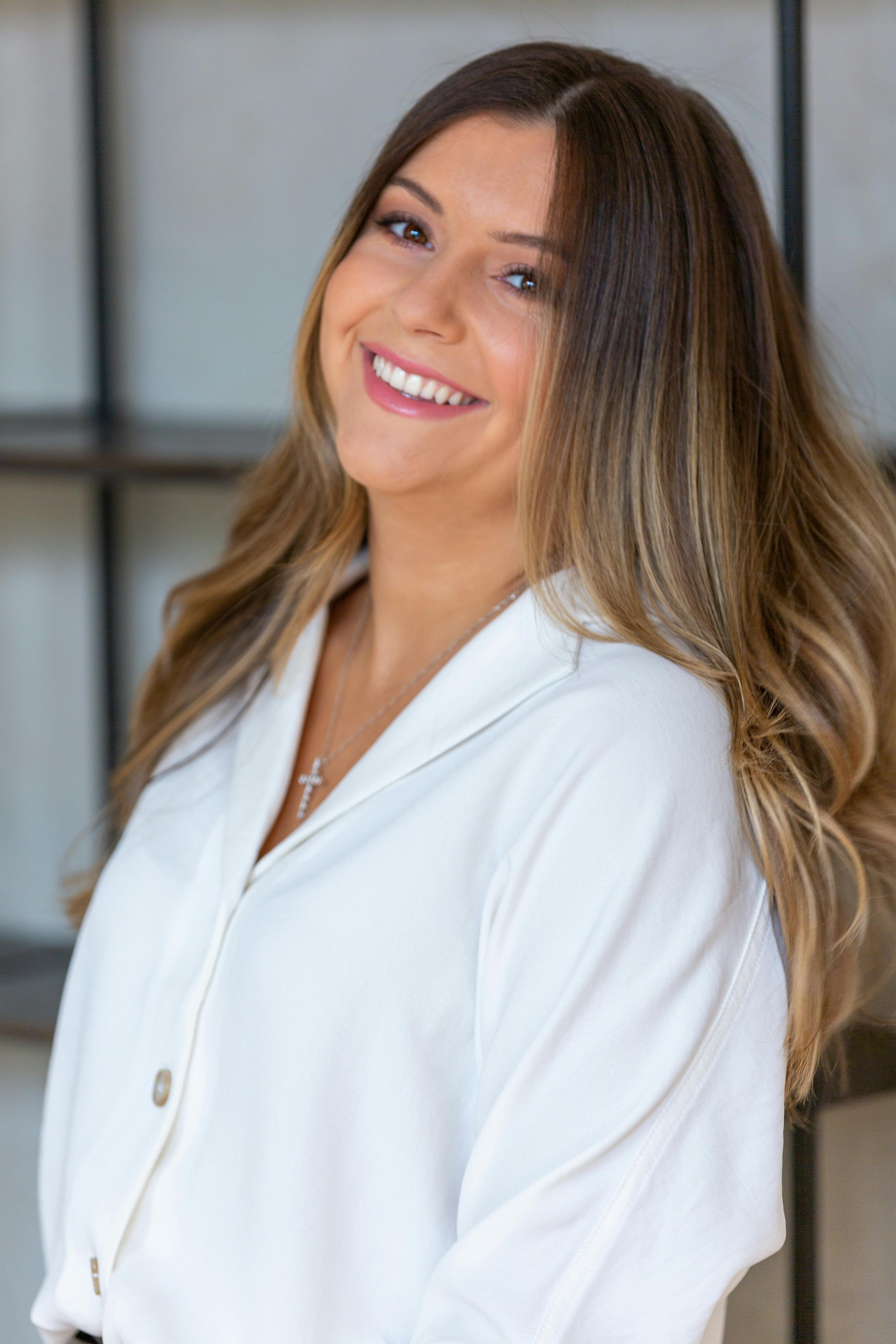Smiling woman in white shirt with a cross necklace, standing indoors. Fashion portrait with natural lighting.