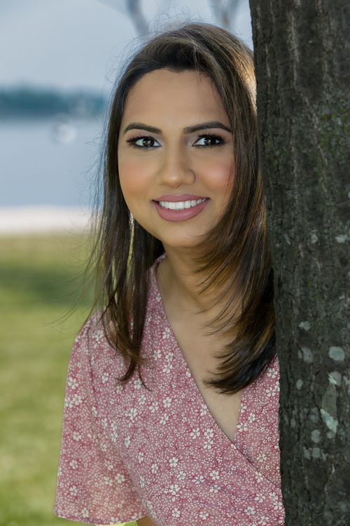 Free A smiling woman in a floral dress leans against a tree outdoors in a sunny setting. Stock Photo