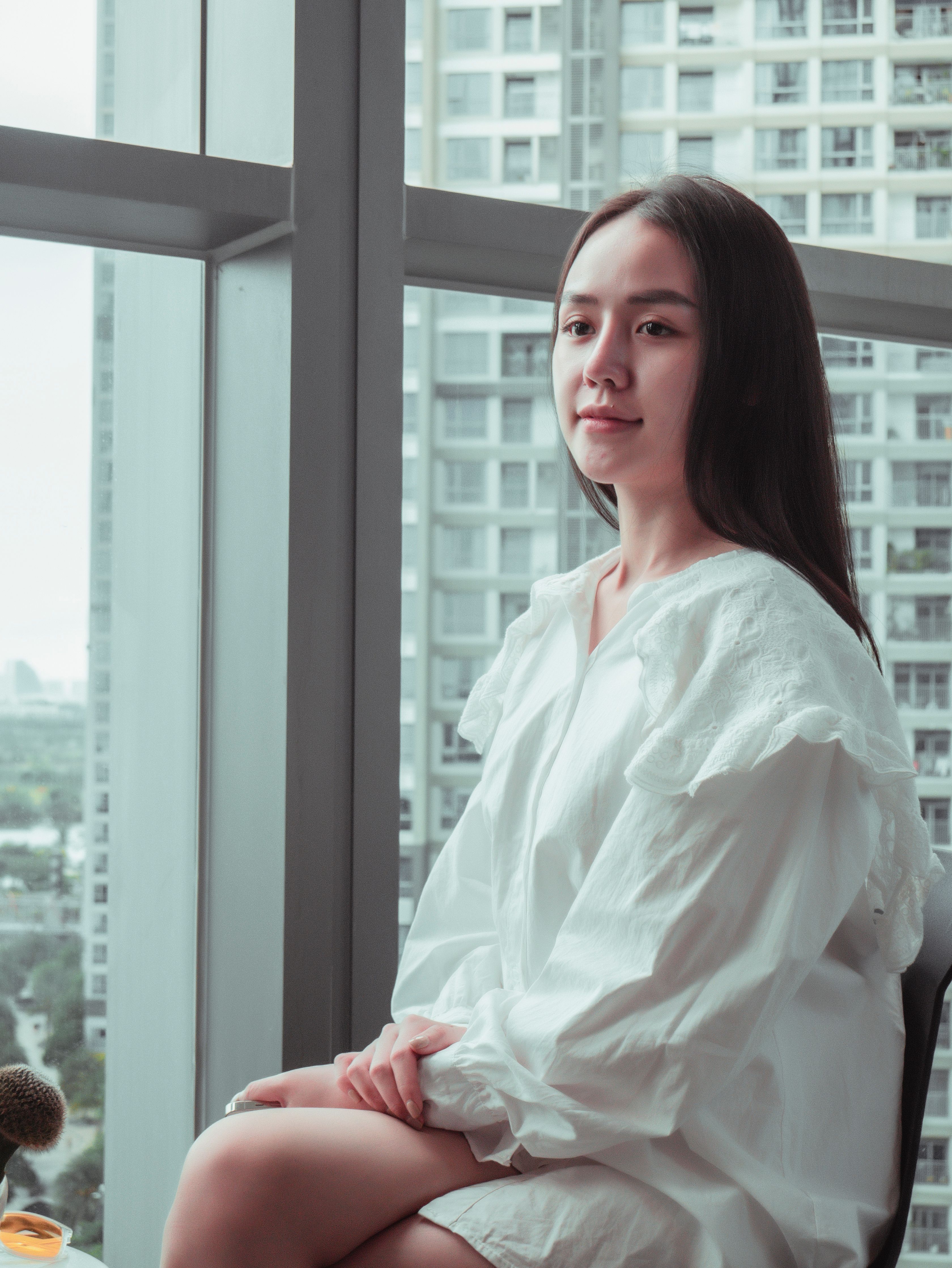 Asian woman in white dress sitting by a window with city view in Vietnam.