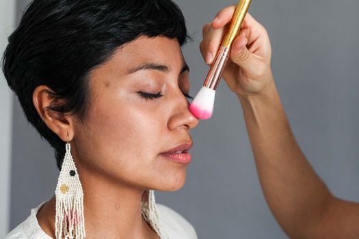 A woman with closed eyes applying makeup with a brush on her nose, showcasing beauty routines.