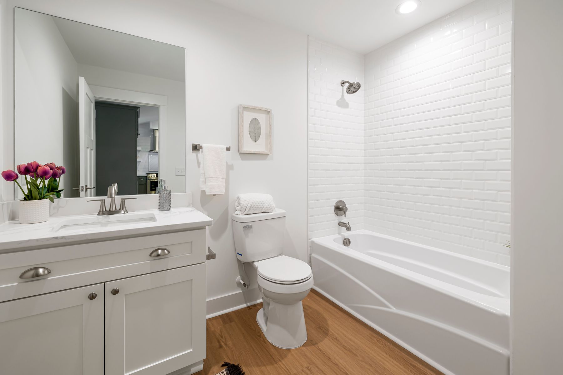 Bright bathroom with broad white surfaces, a bathtub, vanity and warm wood floor