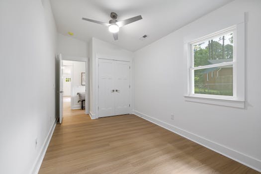 A spacious minimalist empty room with wooden flooring and a large window letting in natural light.