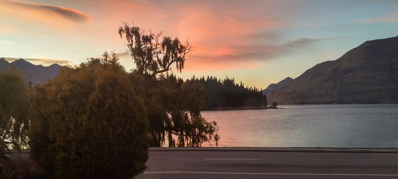Beautiful sunset view over Lake Wakatipu with silhouetted trees and mountains in Queenstown, New Zealand.