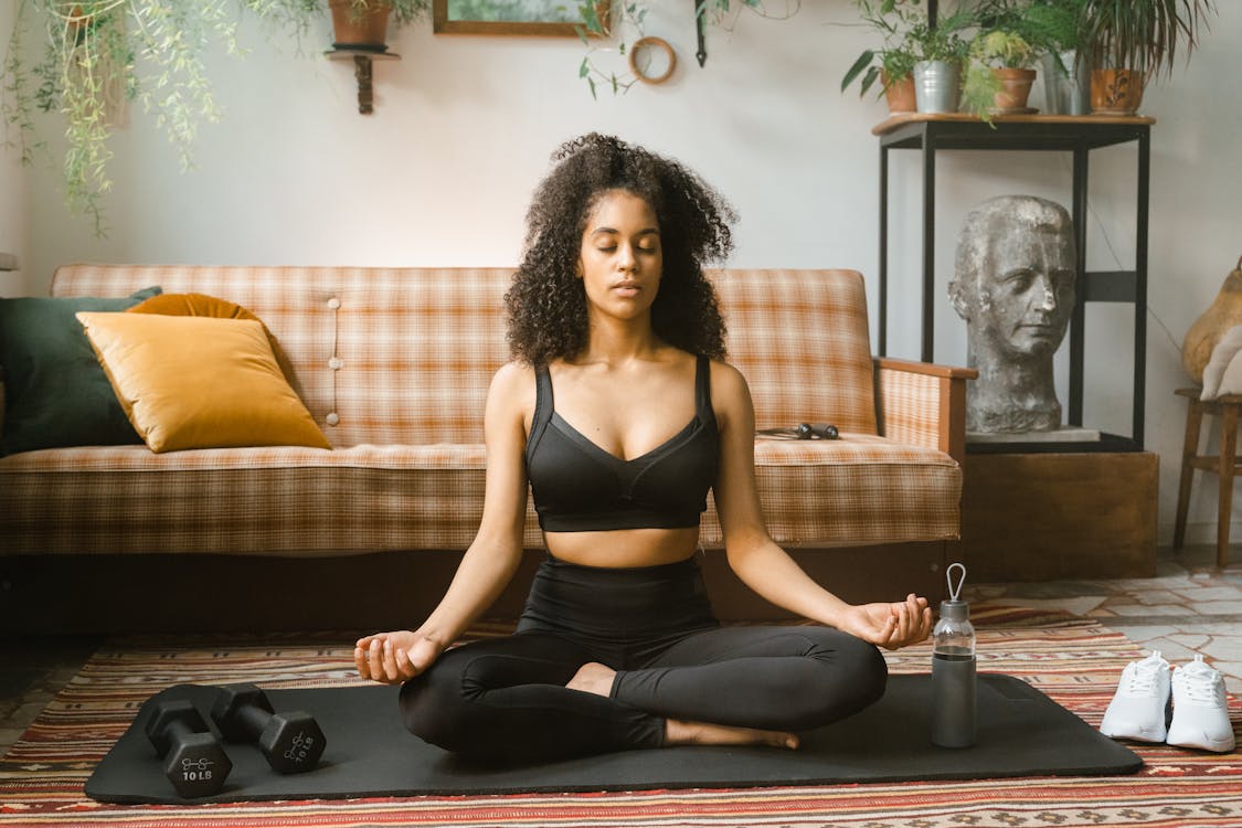 A woman in a black sports bra and yoga pants meditates peacefully in her living room, embodying tranquility and focus.