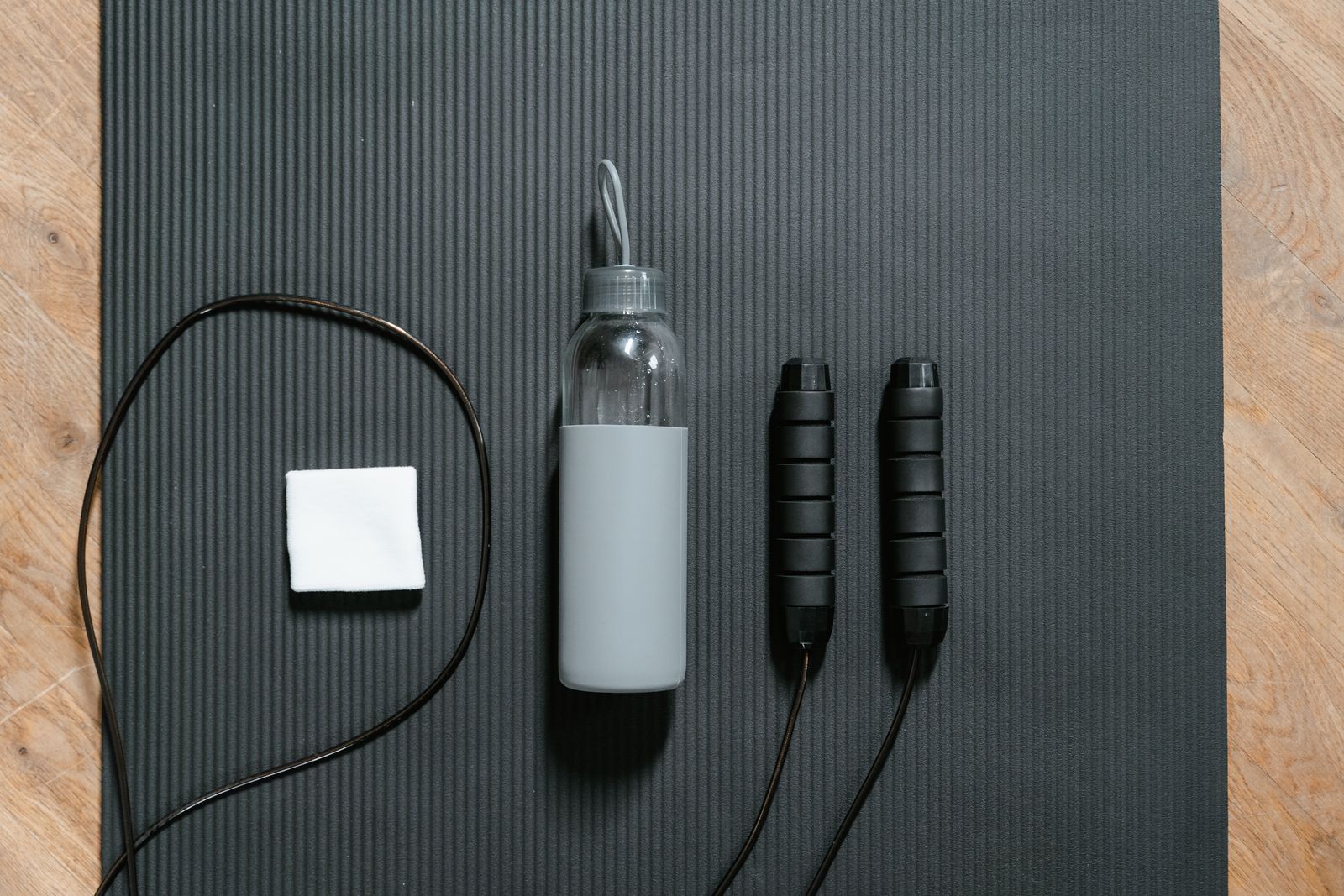 Wide photograph of own-brand goods laid flat: a felt sauna hat, an embroidered cap, a stone gua sha tool, a leather headcover and a laser-engraved steel bottle, on a slate grey surface in even daylight.