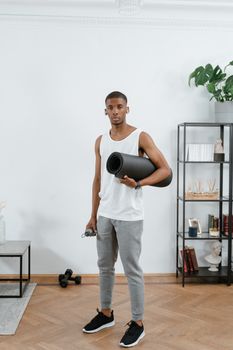 Young man holding yoga mat in a modern home gym setting, ready for workout.