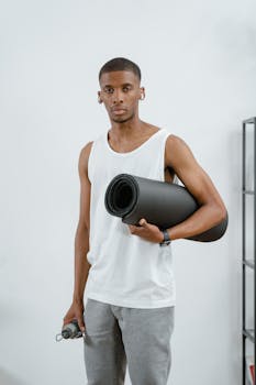A fit man holding a yoga mat and water bottle, ready for a workout at home.