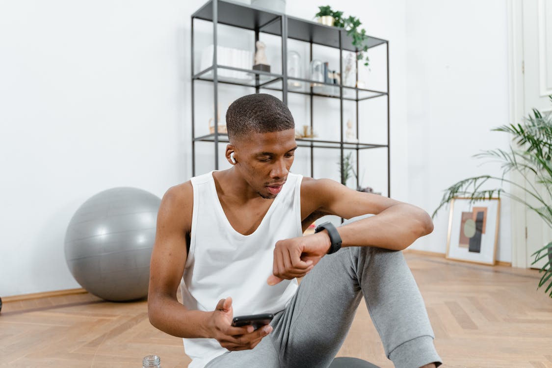  Man Sitting in White Tank Top Looking on His Watch and Holding Smartphone 
