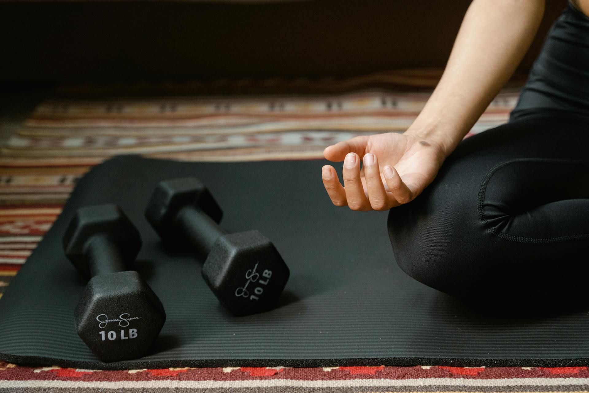 Close-up of a person's hand meditating next to dumbbells on a yoga mat, symbolizing fitness and mindfulness.