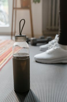 Close-up of a water bottle on a yoga mat with blurred gym equipment in a home setting.