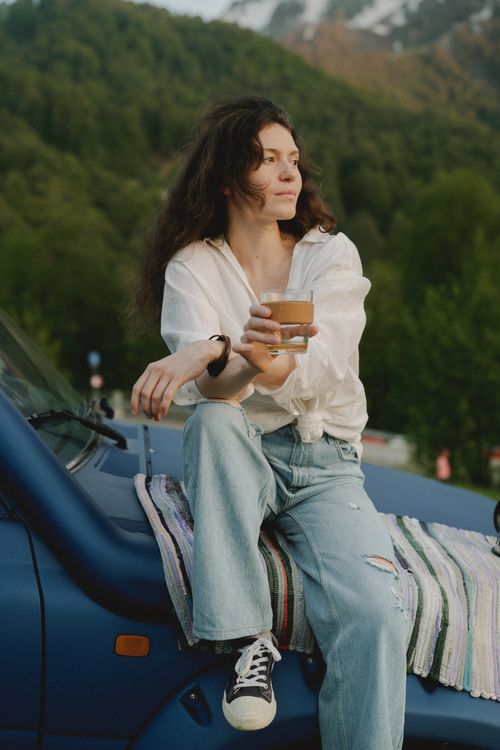 Free Young woman in casual attire sipping a drink while sitting on a car hood in a scenic mountain setting. Stock Photo