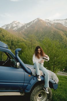 Woman sitting on SUV hood in mountain area with outdoor scenery.
