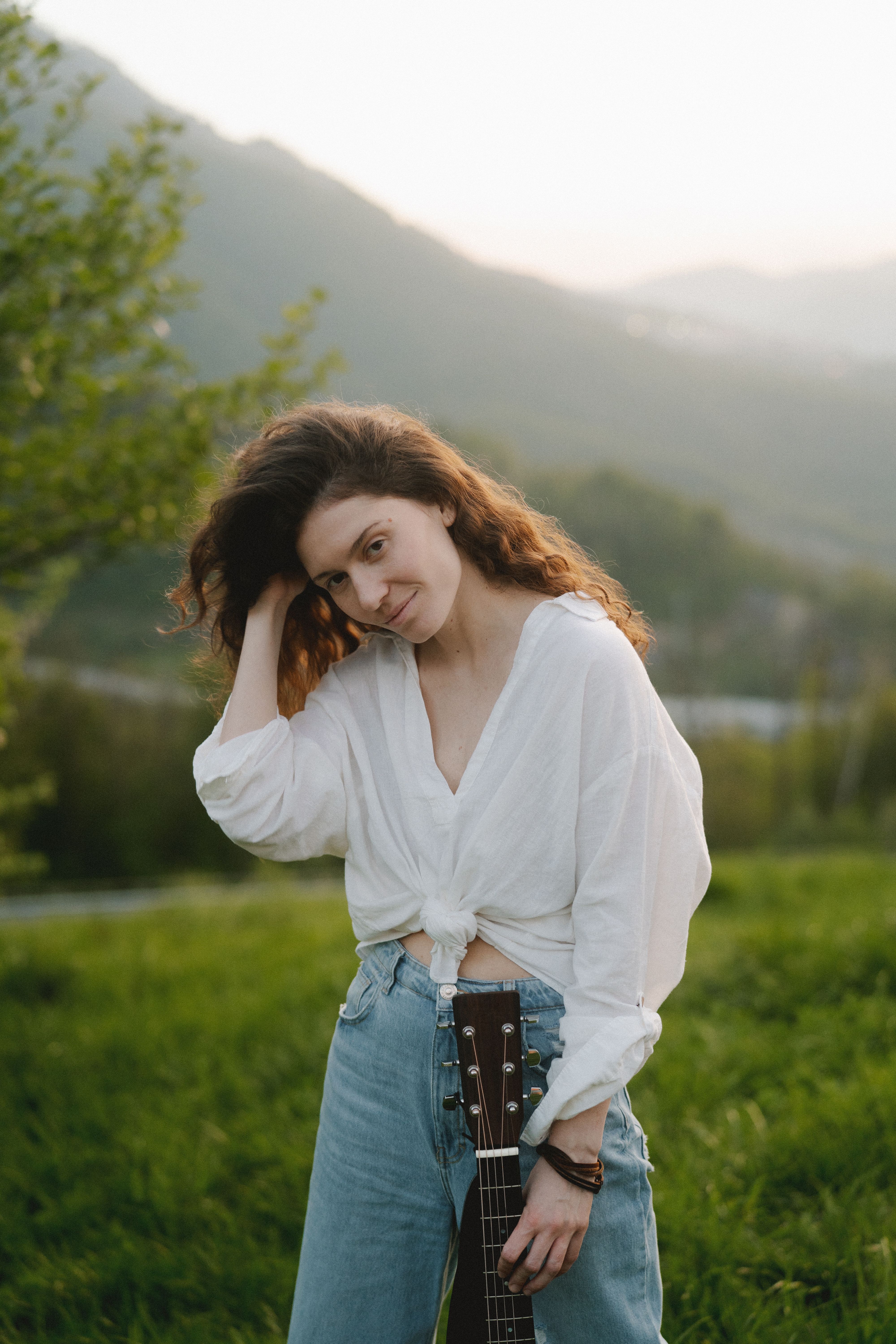 Woman in casual attire posing outdoors with a guitar during sunset.