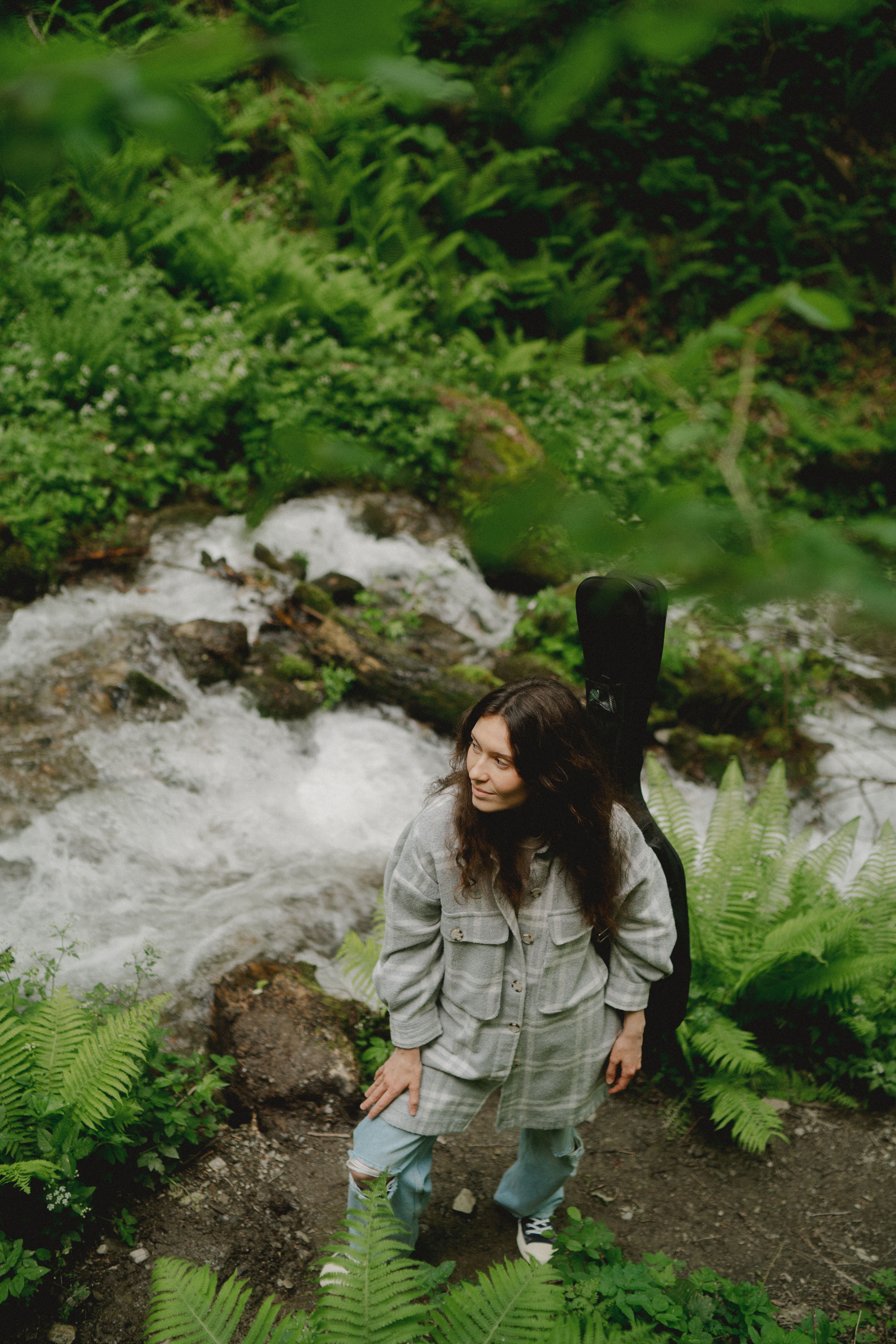 A young woman stands beside a flowing stream in a lush, green forest setting.