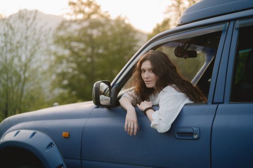 Free Woman in a car enjoying a serene outdoor view with a backdrop of trees and mountains. Stock Photo