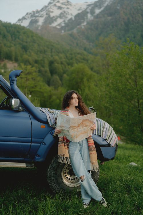 Free A woman leans on a blue car in a mountain scene, reading a large map outdoors. Stock Photo